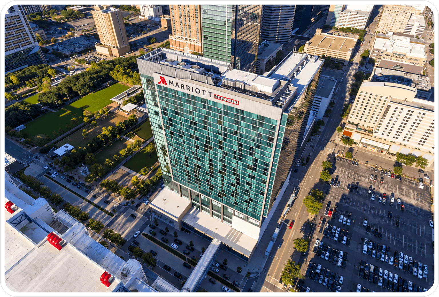 Aerial view of a tall, glass-fronted Marriott Marquis hotel building in an urban area with streets, parked cars, and green spaces surrounding it.